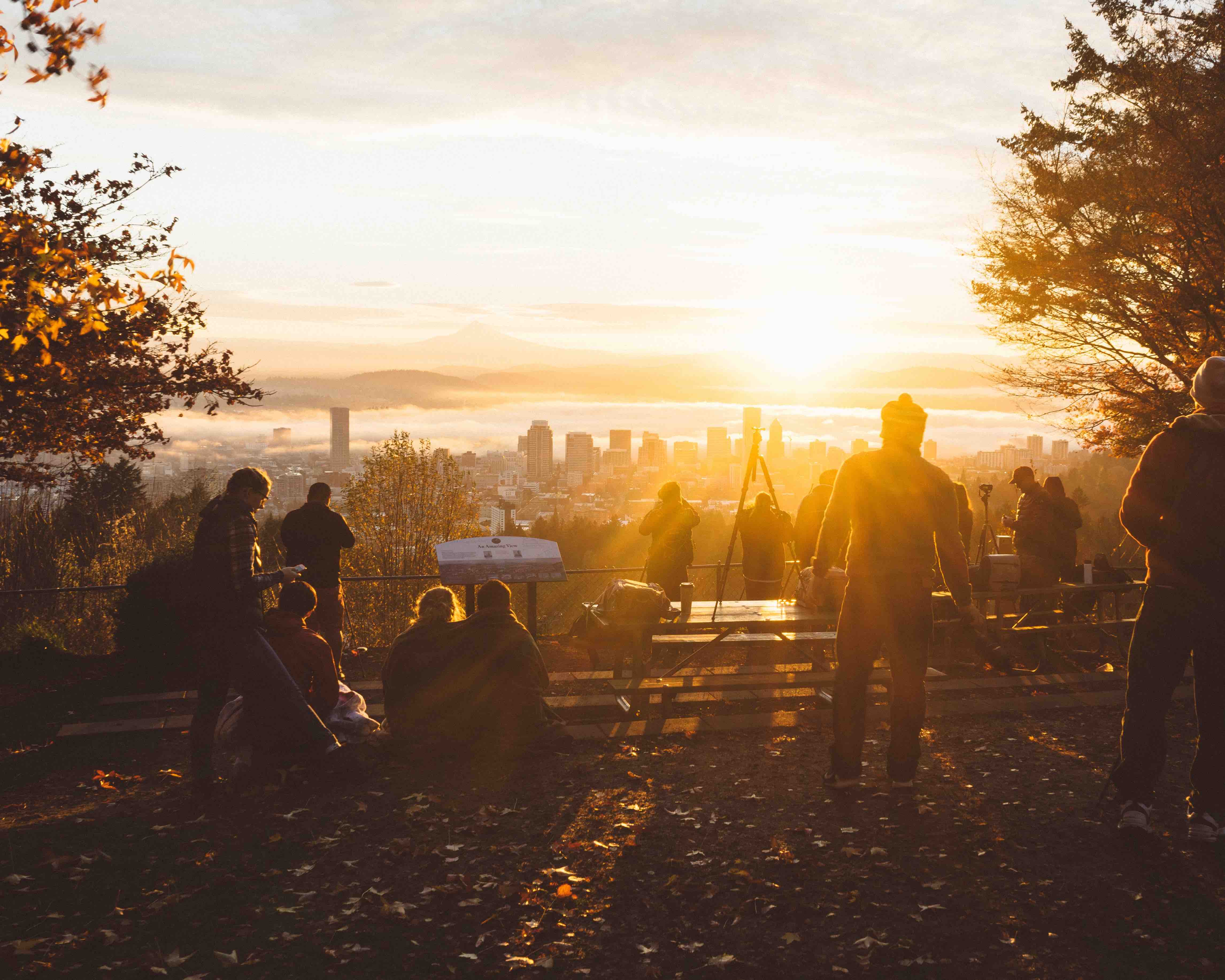 People gathered at a scenic city overlook during sunset, with the skyline and golden sunlight in the background.