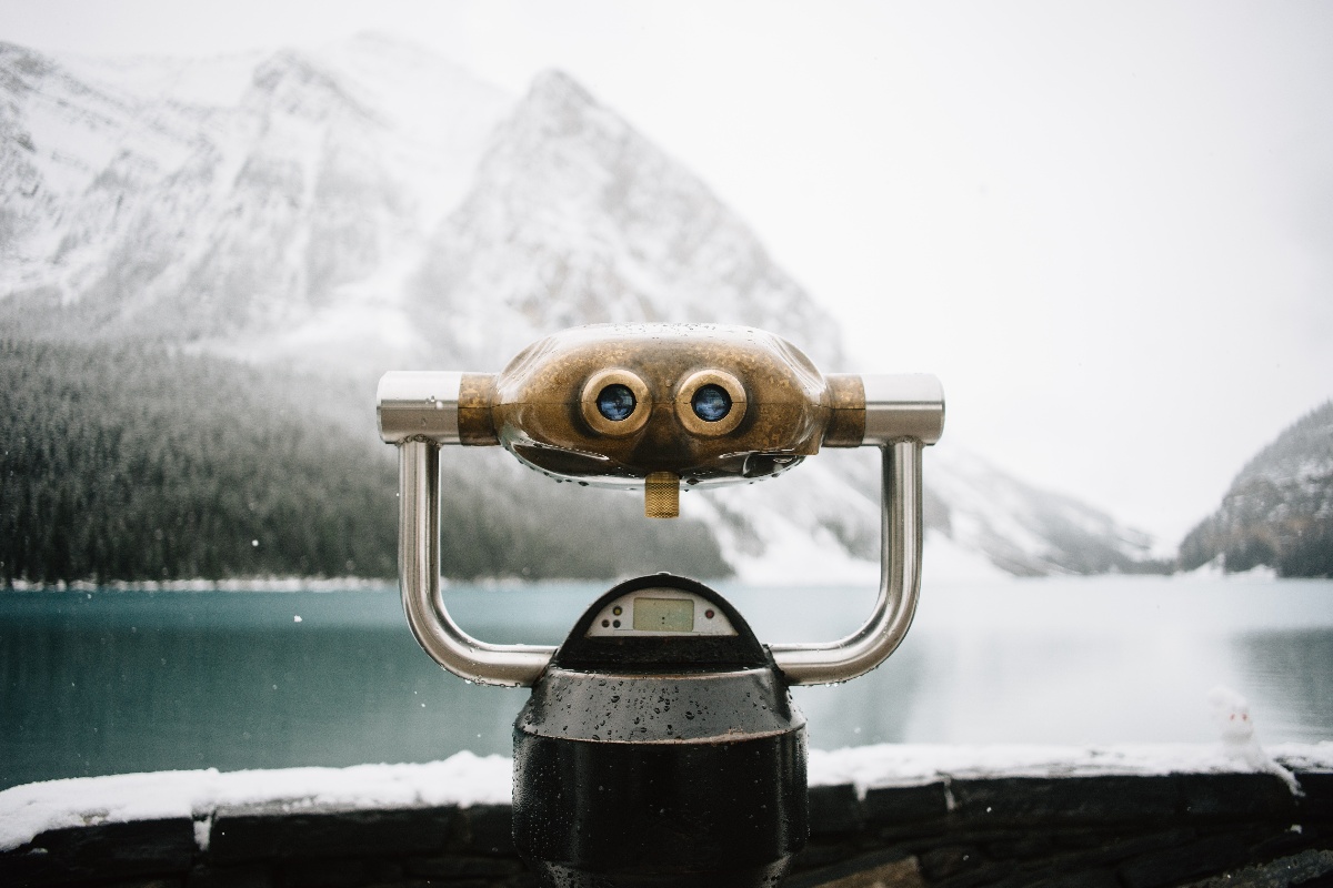 Coin-operated binocular viewer overlooking a snowy mountain lake surrounded by forested peaks.