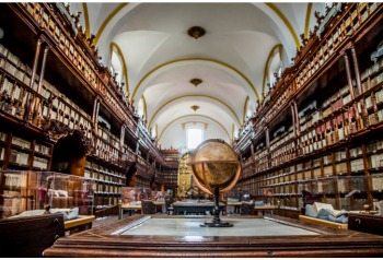 Historic library reading room with floor-to-ceiling bookshelves, long wooden tables and a large globe underneath an arched ceiling