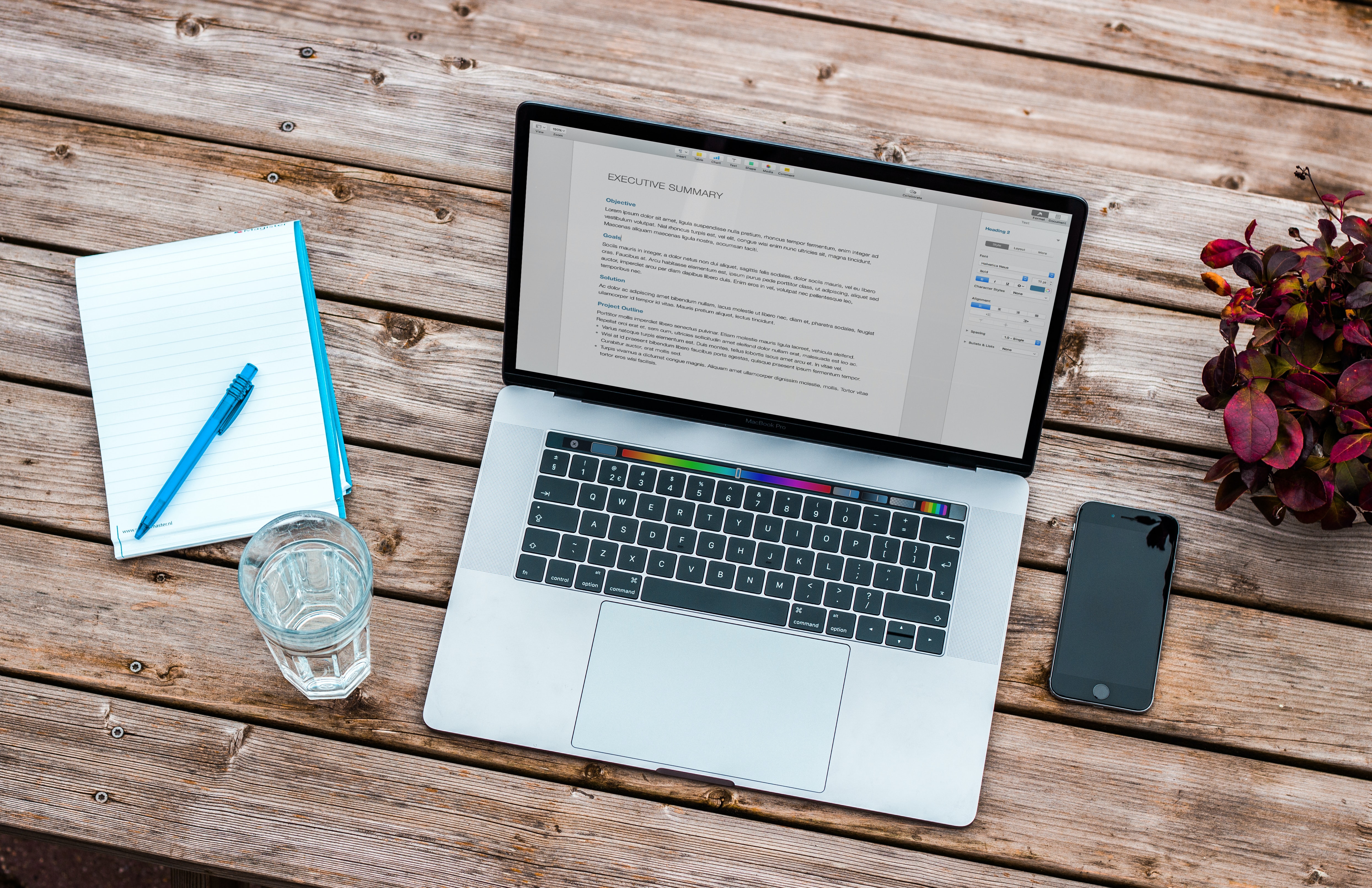 Laptop displaying a document editor on a wooden desk with a notebook, smartphone, and glass of water.