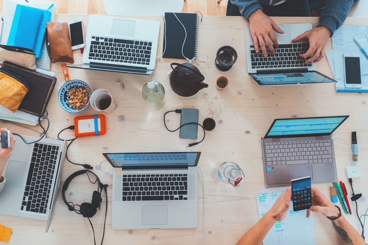 Overhead view of a collaborative workspace with multiple laptops, mobile devices, notebooks, and team members working together.