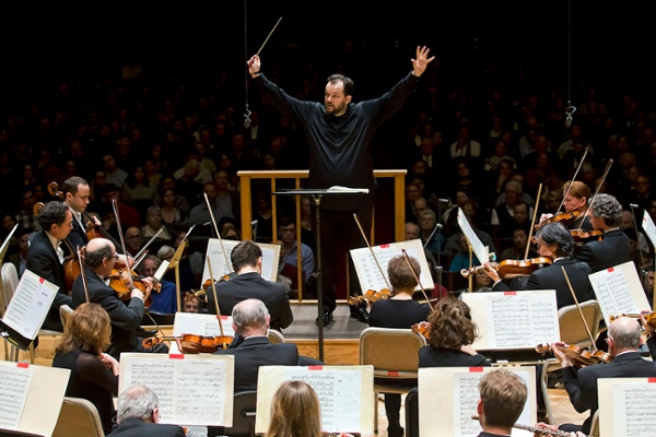 Orchestra conductor leading musicians during a Boston Symphony Orchestra performance, with string players and an audience visible.