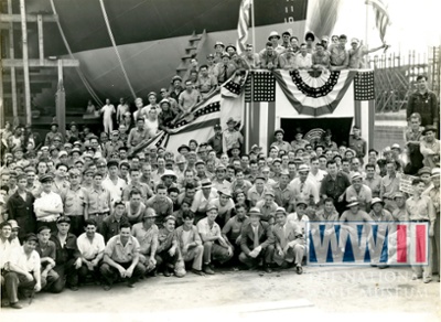 Archival black-and-white photograph of a large group of workers gathered beneath American flags at a World War II-era shipyard.