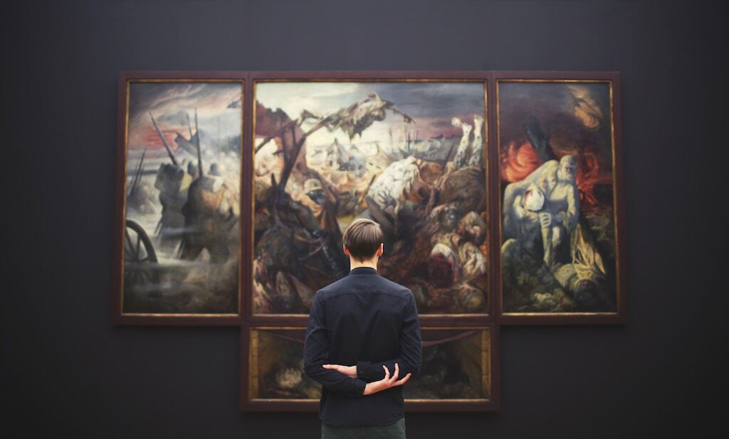 Museum visitor standing in front of a large, three-panel historical painting in a dark art gallery.