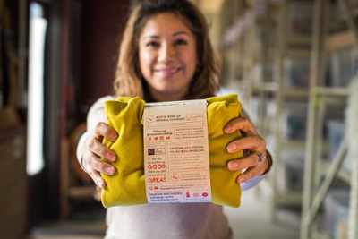 Woman holding a folded yellow garment with The Renewal Workshop label promoting recycled and renewed clothing.