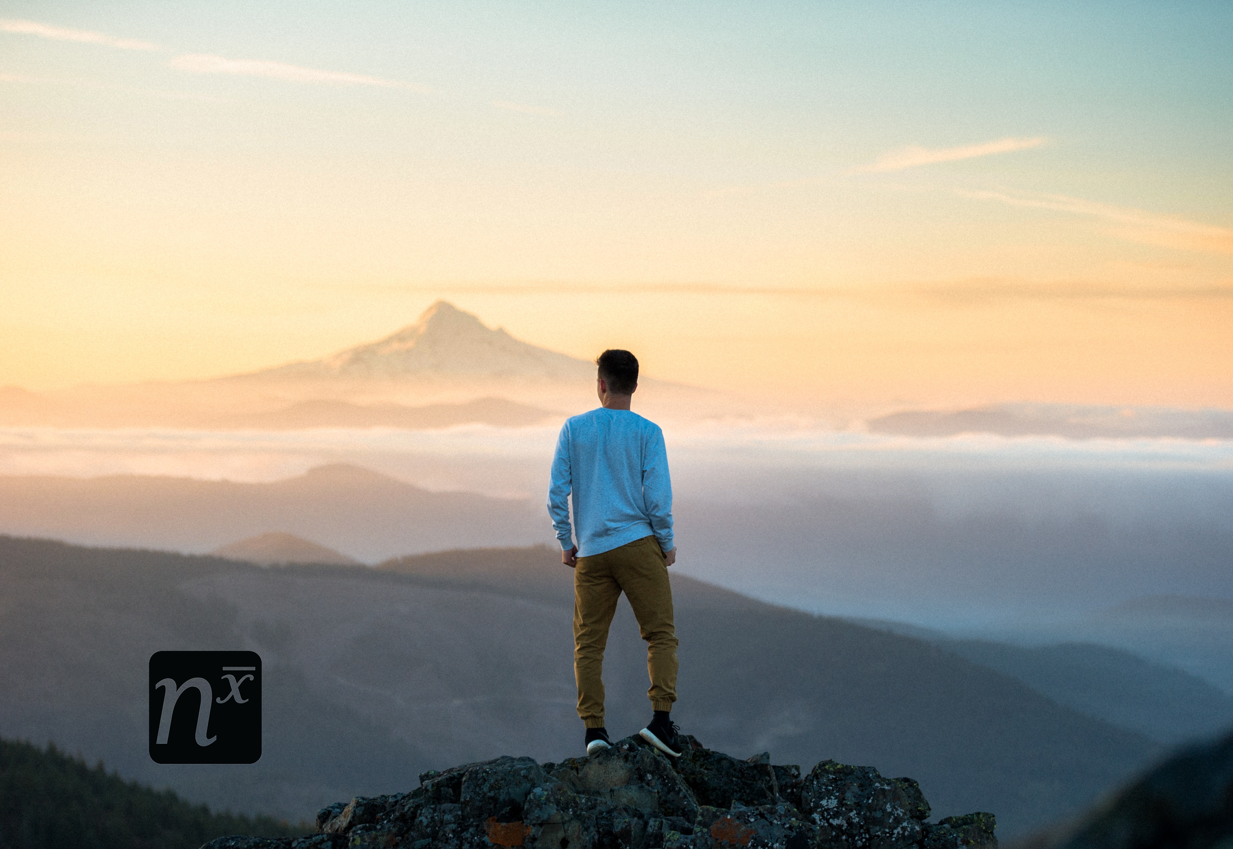 Person standing on a rocky mountain overlook at sunset, with misty valleys and distant peaks in the background.